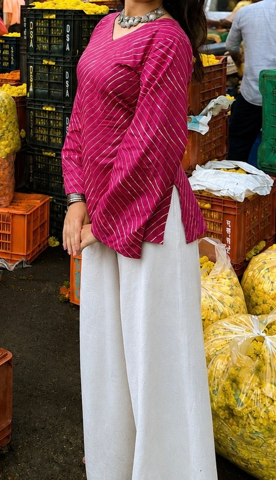 A woman wearing a magenta pink striped long-sleeve blouse and white wide-leg pants, standing in a market setting with crates and bags of yellow produce in the background.